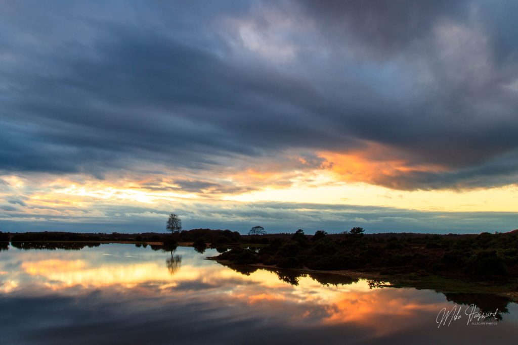 Cloud covers the sunset and produces darks and oranges reflected in the calm water surface of the pond